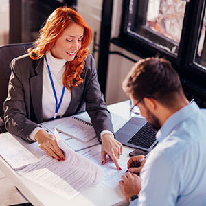 A real estate agent with red hair is pointing at a contract while a man signs it  