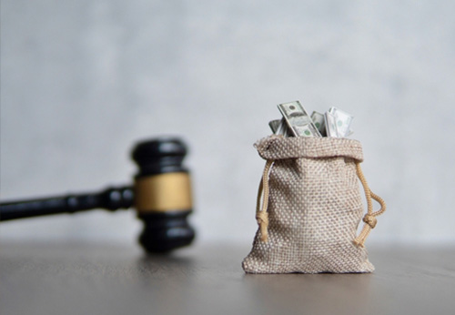 A wooden judge’s gavel resting on a table next to a small burlap sack filled with hundred-dollar bills, symbolizing a court settlement.