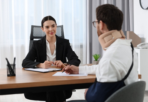 A female attorney in a black suit discussing a case with a male client wearing a neck brace and arm sling in a professional office.
